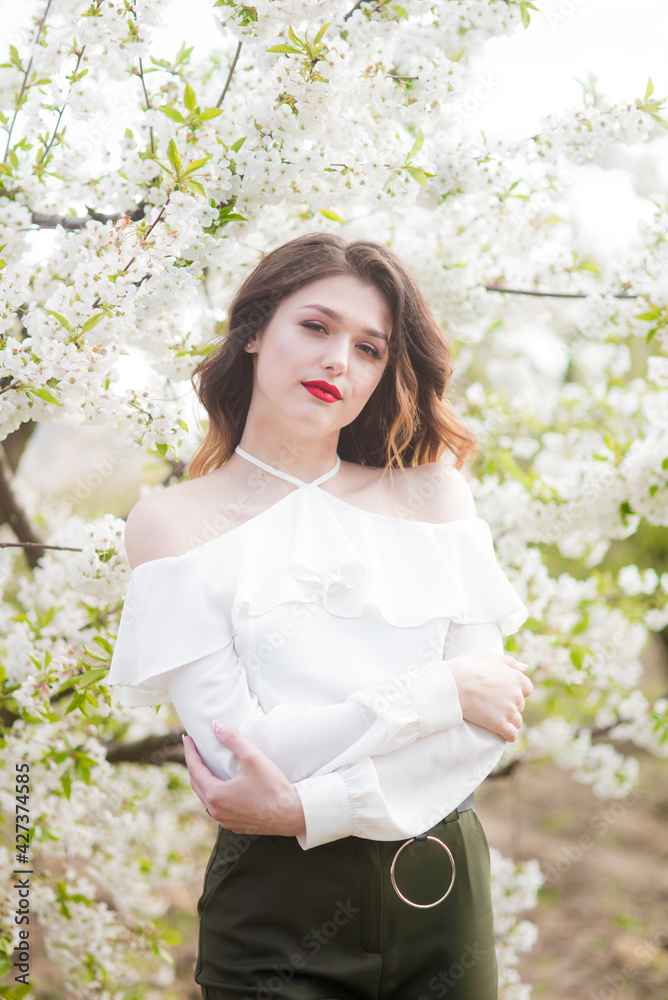 Lovely young girl in a white romantic silk blouse in spring in a blooming cherry garden in sunny weather. Spring and Fashion. Spring blooming gardens Lovely young girl in a white romantic silk blouse in spring in a blooming cherry garden in sunny weather. Spring and Fashion. Spring blooming gardens