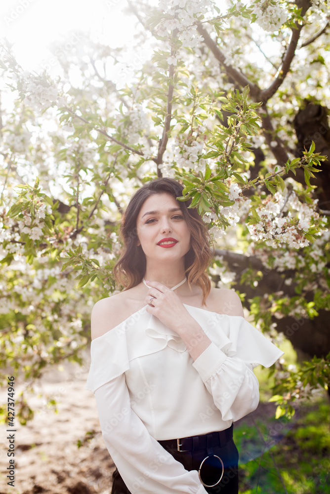 Lovely young girl in a white romantic silk blouse in spring in a blooming cherry garden in sunny weather. Spring and Fashion. Spring blooming gardens Lovely young girl in a white romantic silk blouse in spring in a blooming cherry garden in sunny weather. Spring and Fashion. Spring blooming gardens