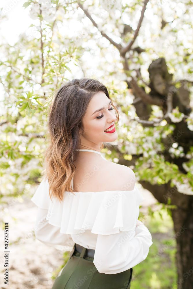 Lovely young girl in a white romantic silk blouse in spring in a blooming cherry garden in sunny weather. Spring and Fashion. Spring blooming gardens Lovely young girl in a white romantic silk blouse in spring in a blooming cherry garden in sunny weather. Spring and Fashion. Spring blooming gardens