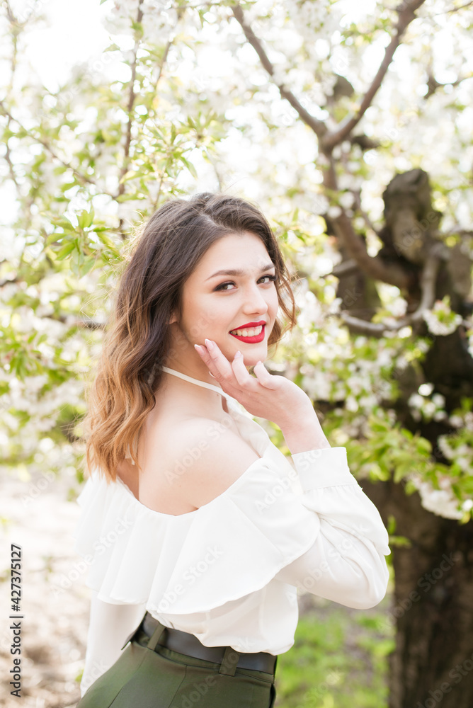 Lovely young girl in a white romantic silk blouse in spring in a blooming cherry garden in sunny weather. Spring and Fashion. Spring blooming gardens Lovely young girl in a white romantic silk blouse in spring in a blooming cherry garden in sunny weather. Spring and Fashion. Spring blooming gardens