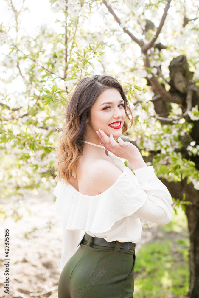 Lovely young girl in a white romantic silk blouse in spring in a blooming cherry garden in sunny weather. Spring and Fashion. Spring blooming gardens Lovely young girl in a white romantic silk blouse in spring in a blooming cherry garden in sunny weather. Spring and Fashion. Spring blooming gardens