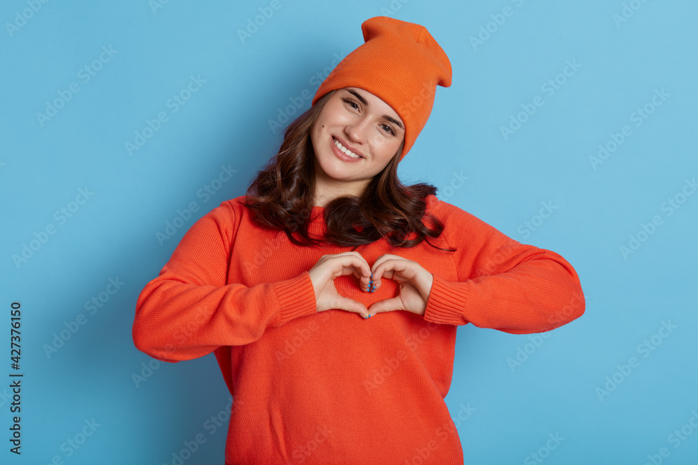Young Caucasian female wearing orange sweater and hat making heart gesture with hands and looking directly at camera with happy expression, isolated over blue background. Young Caucasian female wearing orange sweater and hat making heart gesture with hands and looking directly at camera with happy expression, isolated over blue background.