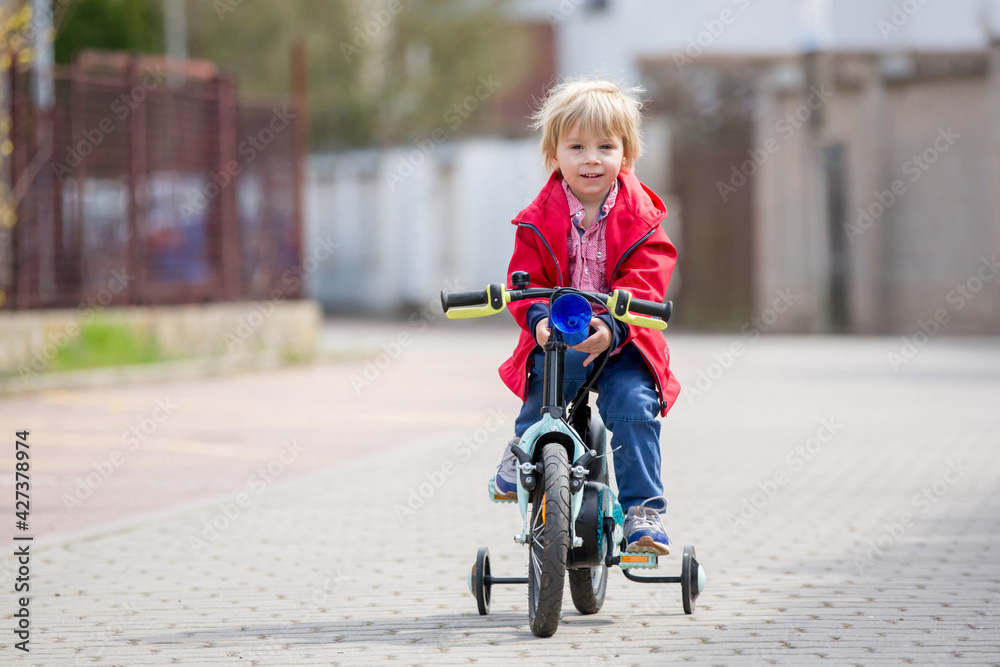 Little child, blond boy, learning how to ride a bicycle in the park using helping wheels on the side Little child, blond boy, learning how to ride a bicycle in the park using helping wheels on the side