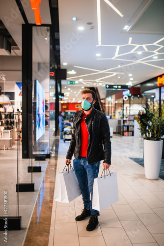 A young bearded Caucasian man in a black leather jacket, with a mask and bags in his hand stands next to a shop window in a shopping center during the COVID-19 coronavirus epidemic