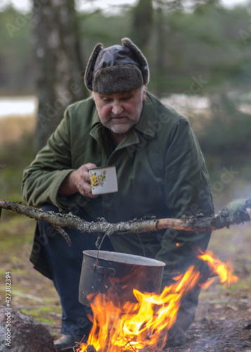 a forester in the forest drinks tea at rest, blurred forest background, bonfire with a pot over it, in the autumn