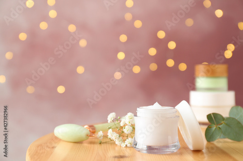 Natural face roller, cosmetic product and beautiful flowers on wooden table against blurred lights, space for text