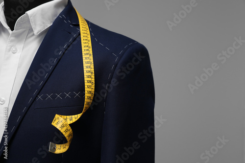 Semi-ready jacket with tailor's measuring tape on mannequin against grey background, closeup. Space for text photo