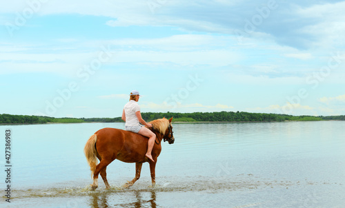 One Caucasian woman is riding her red horse along the water in a shallow lagoon.