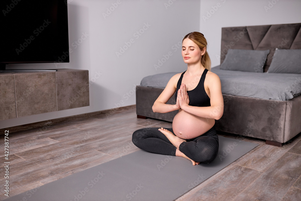 Fototapeta premium side view on calm pregnant female sitting on floor, keeping hands together, namaste