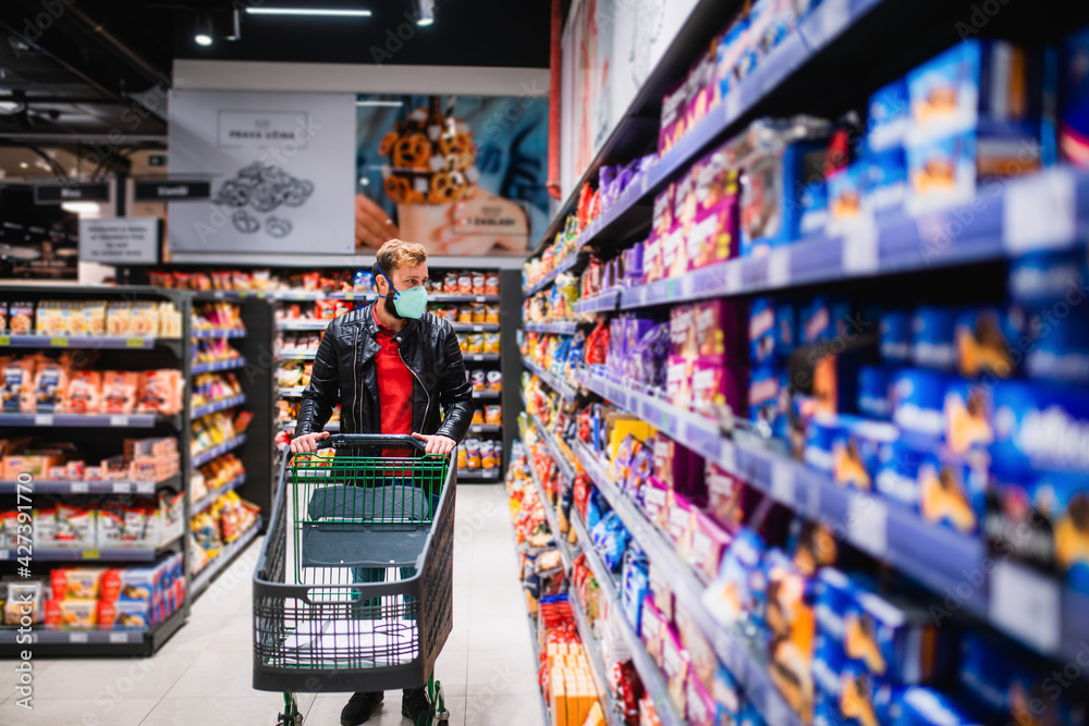 Fototapeta premium A young man with a medical face mask pushes a basket to the supermarket and looks at groceries. Procurement of food during the coronavirus pandemic COVID - 19