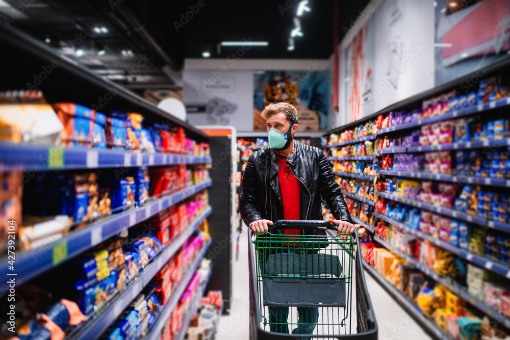 A young handsome man with a medical face mask buys food at the supermarket and pushes a grocery basket. Purchase of food during the coronavirus pandemic COVID - 19