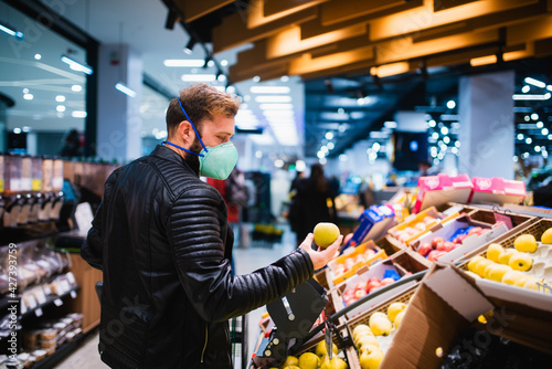 Side view of a man with a face mask holding an apple in his hand and buying healthy groceries at the supermarket. Procurement of food during the coronavirus crisis COVID - 19 pandemic