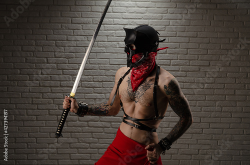 a samurai man in a demon mask with tattoos and a katana sword in his hands against the background of a brick wall