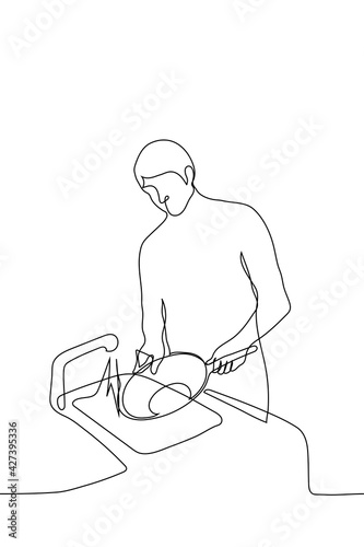 man washing dishes - one line drawing vector. a householder or waiter washes a large frying pan while standing in front of the sink with the tap on