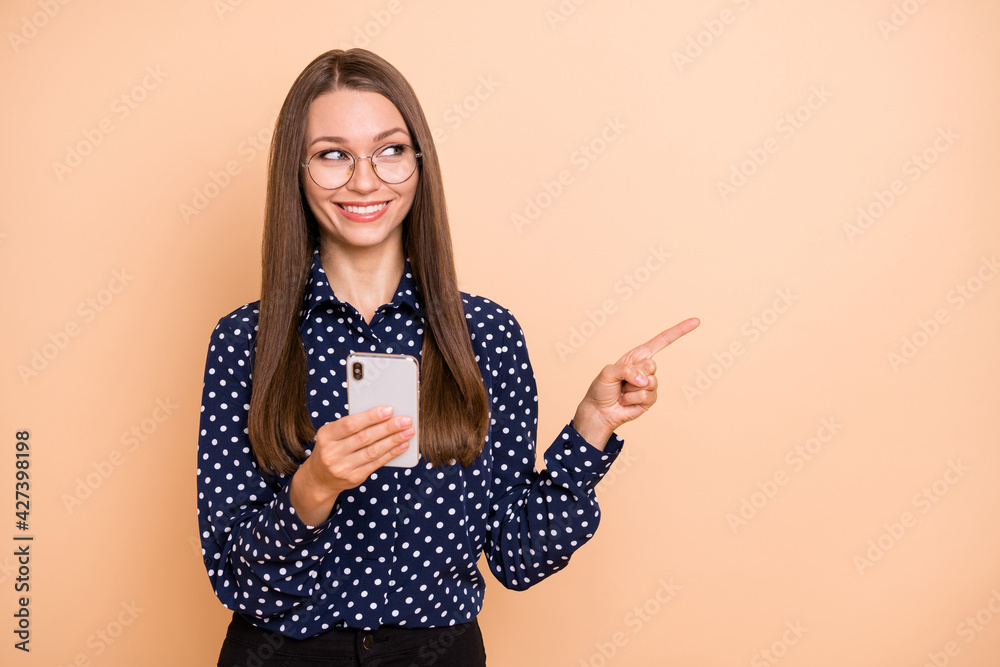 Photo of charming pretty girl dressed dotted blouse glasses reading modern device pointing empty space isolated beige color background Photo of charming pretty girl dressed dotted blouse glasses reading modern device pointing empty space isolated beige color background
