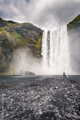 A couple in front of a famous Skogafoss waterfall, Iceland. Cloudy day in Icelandic nature.