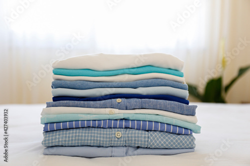 Stack of colorful perfectly folded clothing items on the bed at home. Pile of different pastel color shirts and t-shirts isolated on pale pink background. Close up, copy space.