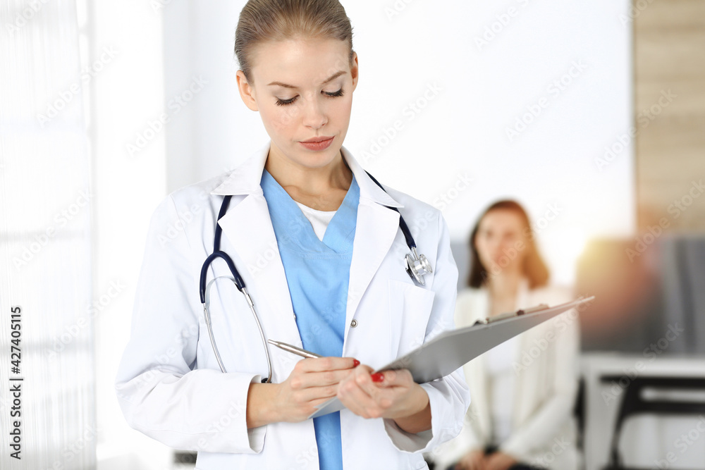 Woman - doctor is using clipboard for filling up medication history record form in sunny clinic at the background of patient Woman - doctor is using clipboard for filling up medication history record form in sunny clinic at the background of patient
