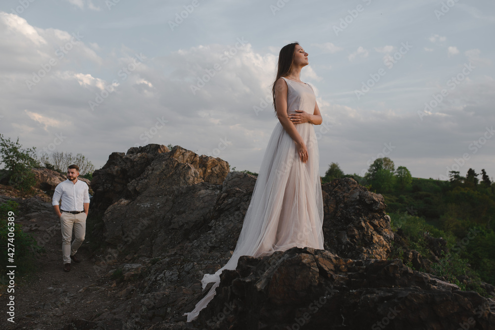 Close up beautiful tender bride woman in long wedding dress standing on the big rock. Groom walking to the bride. Mountains, evening. Close up beautiful tender bride woman in long wedding dress standing on the big rock. Groom walking to the bride. Mountains, evening.