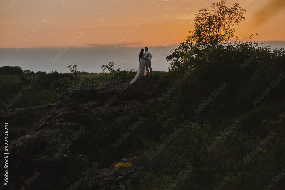 Rear view of romantic wedding couple of groom and bride in gorgeous wedding dress with long loop standing on the rocks in the mountains on sunset. Scenic landscape view