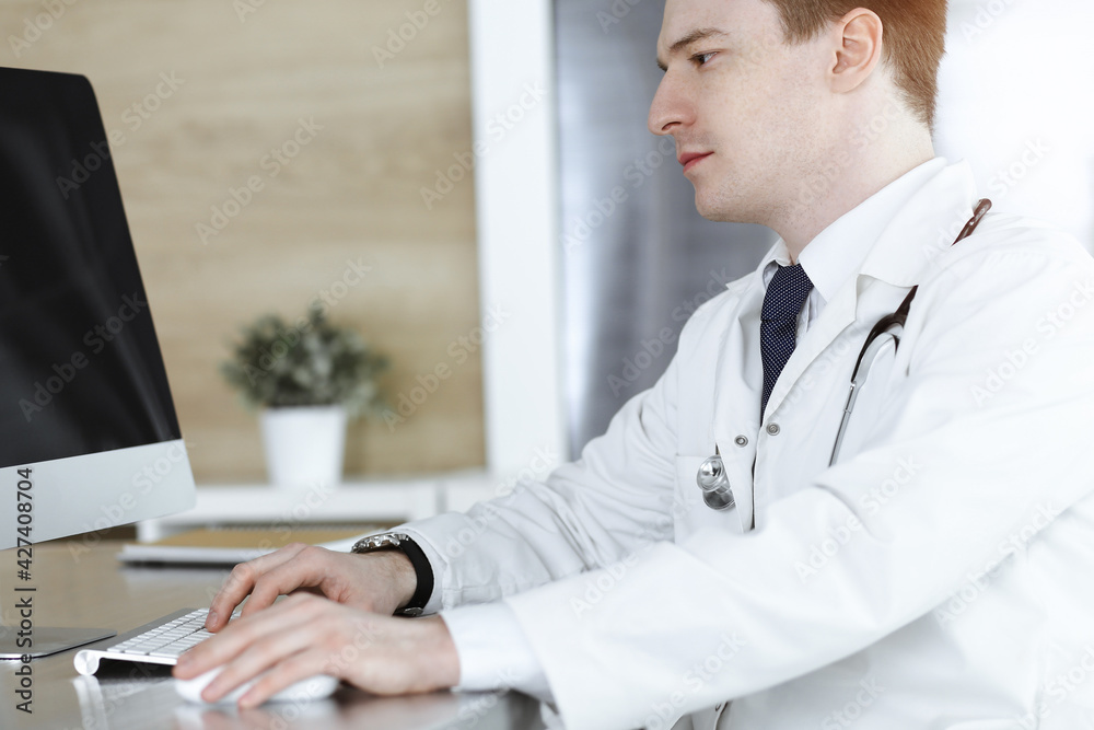 Doctor man sitting behind a computer at workplace in sunny clinic