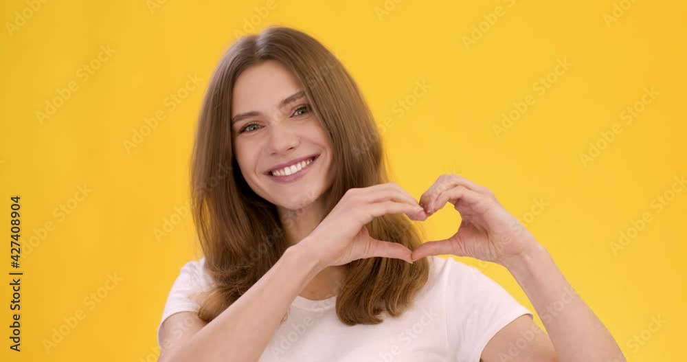 Kindness concept. Young cute positive lady showing heart shape with her hands and smiling to camera, orange background Kindness concept. Young cute positive lady showing heart shape with her hands and smiling to camera, orange background