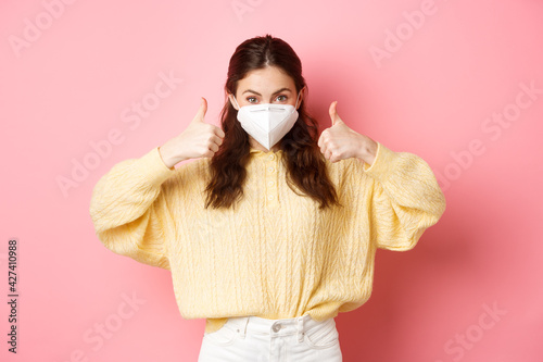 Covid-19, lockdown and pandemic concept. Young woman wearing respirator, face mask during quarantine, showing thumbs up in approval, support vaccination, pink background photo