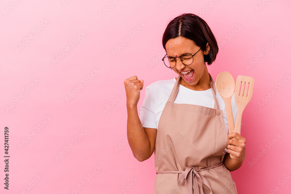 Young indian cook woman holding a wooden spoon and fork isolated raising fist after a victory, winner concept. Young indian cook woman holding a wooden spoon and fork isolated raising fist after a victory, winner concept.