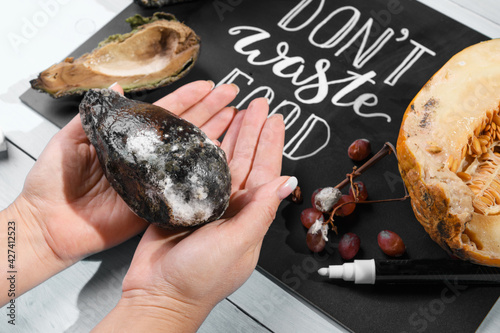 Close up of female hand holding a moldy avocado on a chalkboard with Don't waste food lettering and rotten fruits. Selective focus. Concept of stop wasting food photo