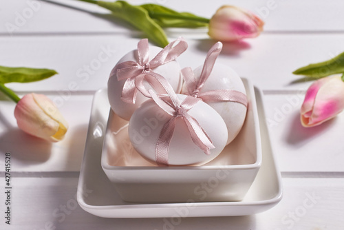 Three white eggs with pink bows on a porcelain plate on a white wooden background and tulip flowers.