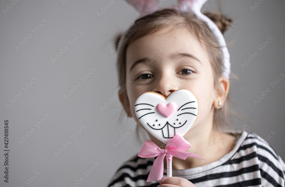 A cute girl with Easter bunny ears on her head holds a gingerbread on a stick.