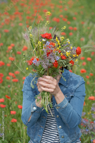 Portrait of young woman in denim jacket on flower meadow background. Woman hides face behind bouquet of beautiful wildflowers. Spring bright flowers backdrop, red poppy field. Country landscape. © Olga