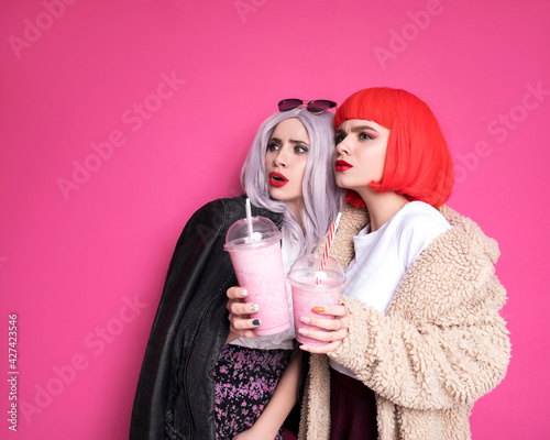 Two cute girl friends woman looking like teenagers standing on a pink background. They wearing colorful wigs and stylish casual outfits and eating pizza with milkshakes. Happy emotional portraits photo