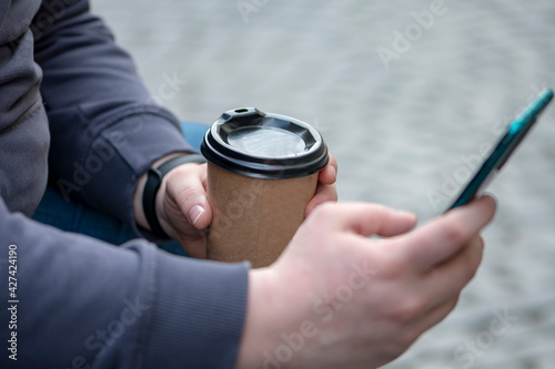 Fotografie The man looks at his mobile phone and holds a coffee in a paper cup in his hand