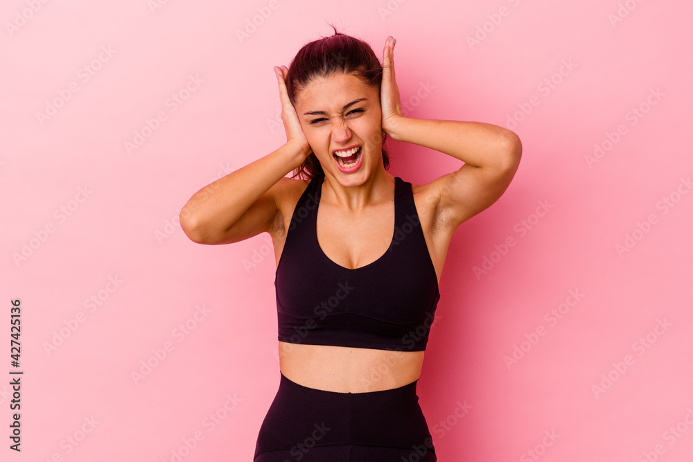 Young sport Indian woman isolated on pink background covering ears with hands trying not to hear too loud sound.