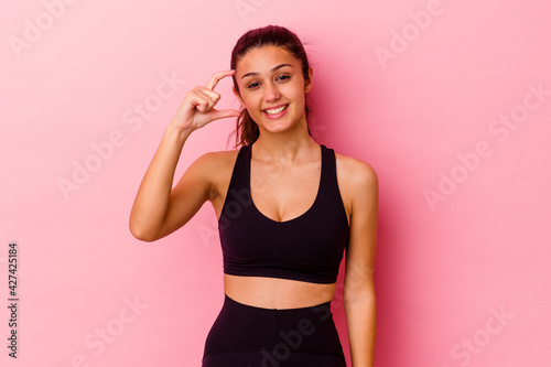 Young sport Indian woman isolated on pink background holding something little with forefingers  smiling and confident.