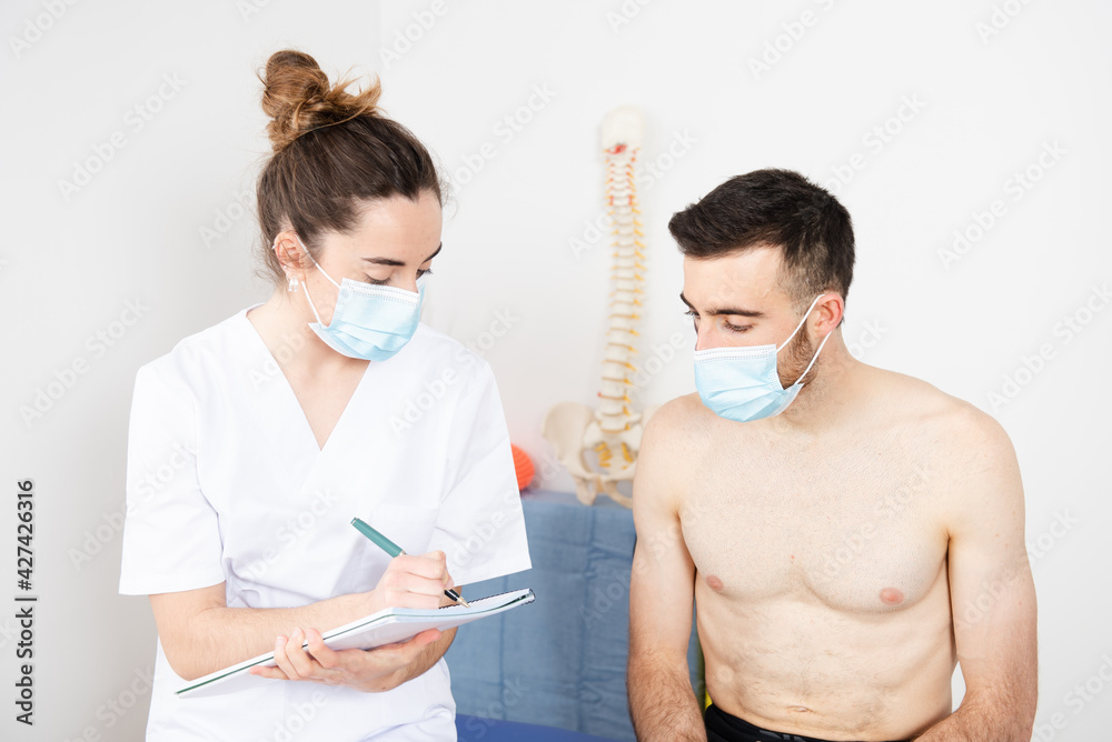 Chiropractor working with a patient and writting on a notebook while wearing face masks in a physiotherapy center during Coronavirus pandemic.
