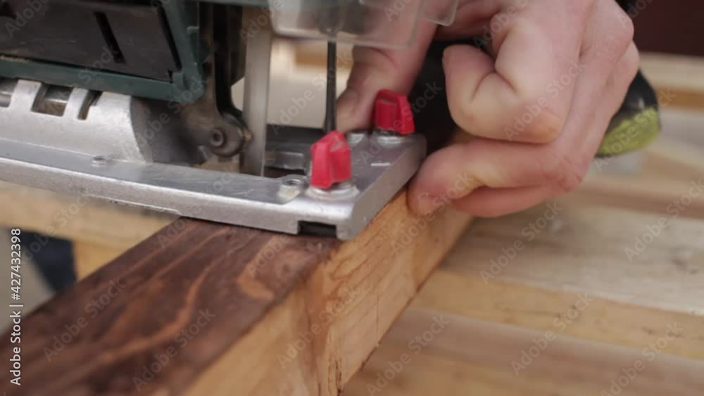 A young man cuts the boards with a fretsaw. Automatic electric saw on wood. Pine beam. The legs for the table with their own hands. Cheap eco-friendly garden furniture top A young man cuts the boards with a fretsaw. Automatic electric saw on wood. Pine beam. The legs for the table with their own hands. Cheap eco-friendly garden furniture top