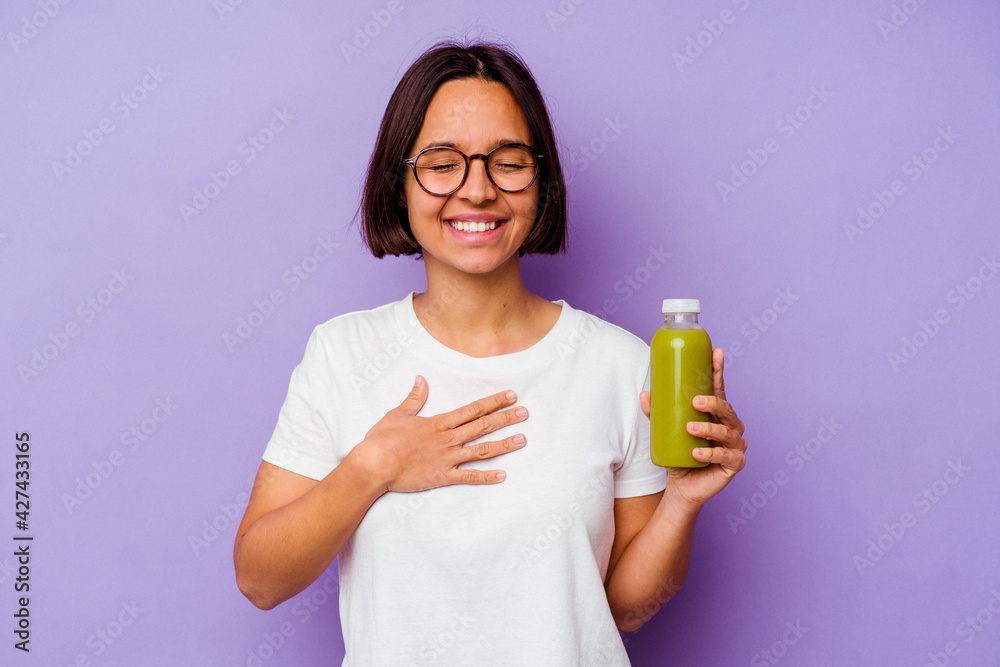 Young mixed race woman holding a healthy smoothie isolated on purple background laughs out loudly keeping hand on chest. Young mixed race woman holding a healthy smoothie isolated on purple background laughs out loudly keeping hand on chest.