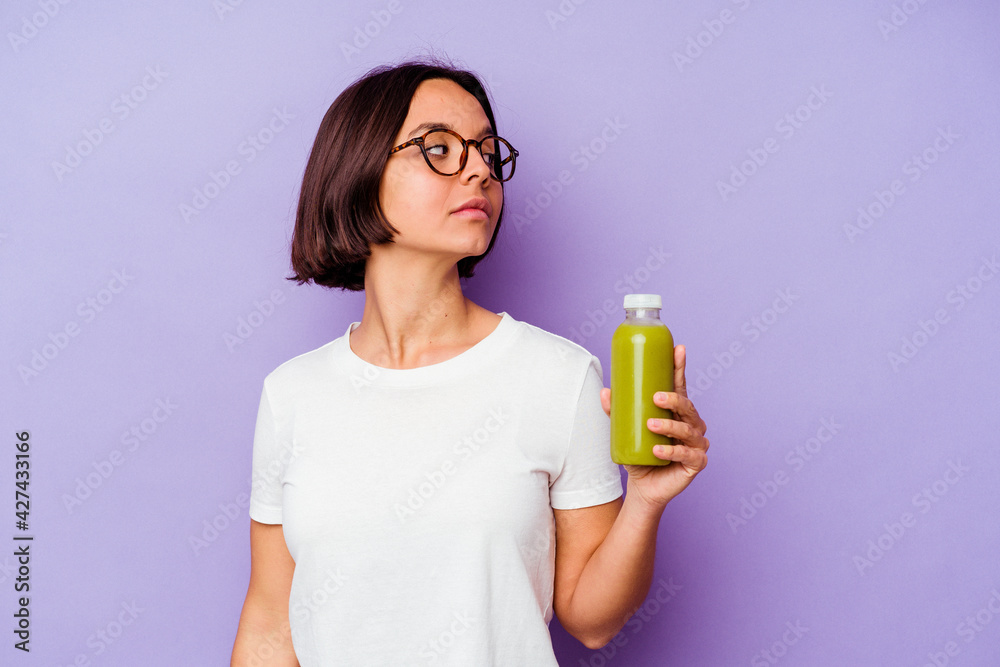 Young mixed race woman holding a healthy smoothie isolated on purple background looks aside smiling, cheerful and pleasant. Young mixed race woman holding a healthy smoothie isolated on purple background looks aside smiling, cheerful and pleasant.
