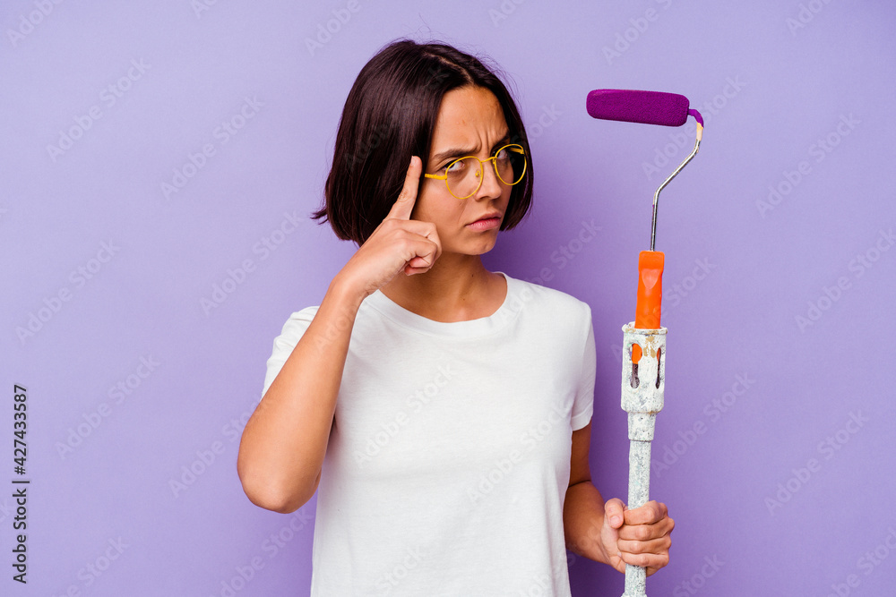 Young painter mixed race woman holding a paint stick isolated on purple background pointing temple with finger, thinking, focused on a task. Young painter mixed race woman holding a paint stick isolated on purple background pointing temple with finger, thinking, focused on a task.