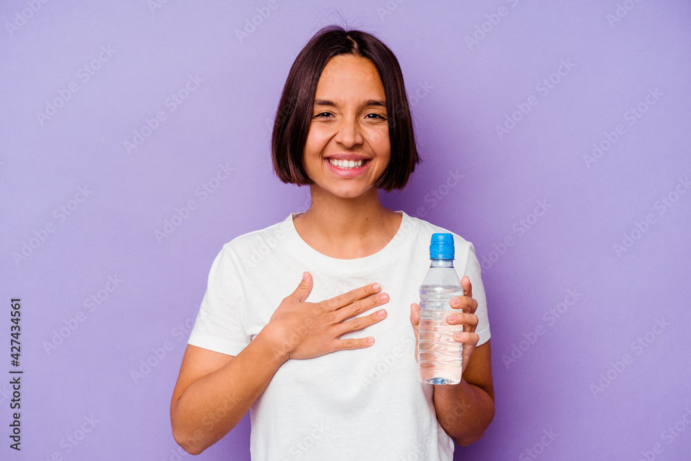 Young mixed race holding a water bottle isolated on purple background laughs out loudly keeping hand on chest. Young mixed race holding a water bottle isolated on purple background laughs out loudly keeping hand on chest.