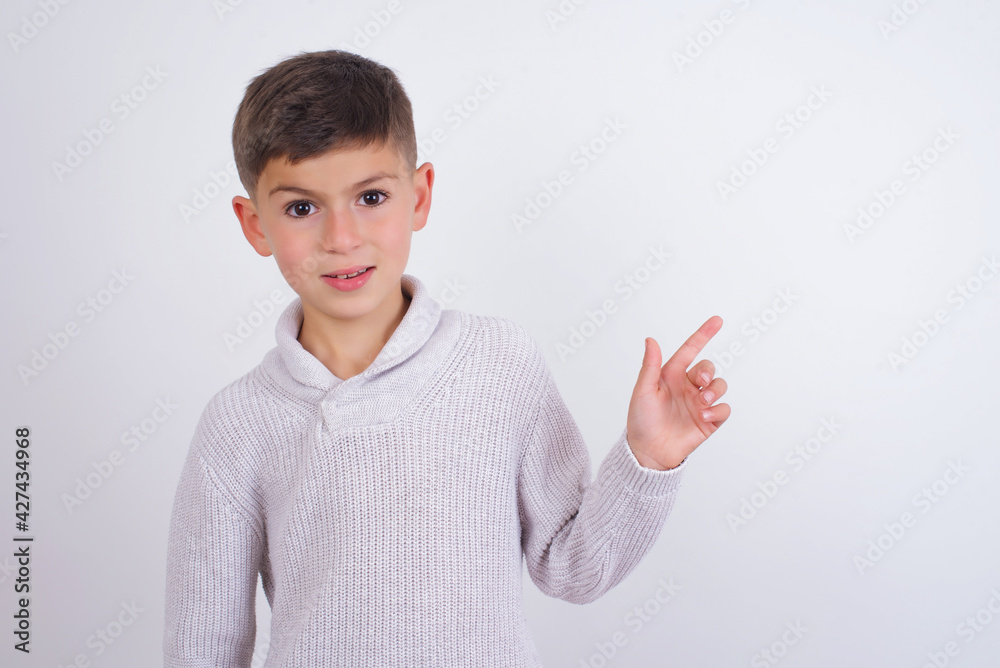 Caucasian kid boy wearing knitted sweater against white wall looking at camera indicating finger empty space sales Caucasian kid boy wearing knitted sweater against white wall looking at camera indicating finger empty space sales