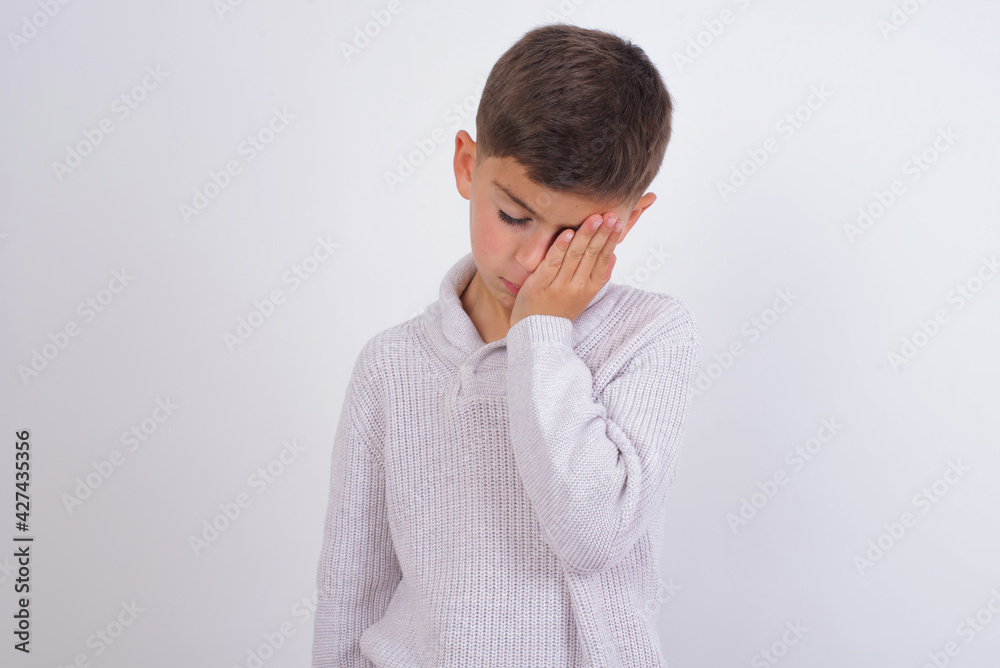 Tired overworked Caucasian kid boy wearing knitted sweater against white wall has sleepy expression, gloomy look, covers face with hand, has eyes shut, gasps from tiredness, fatigue after party Tired overworked Caucasian kid boy wearing knitted sweater against white wall has sleepy expression, gloomy look, covers face with hand, has eyes shut, gasps from tiredness, fatigue after party