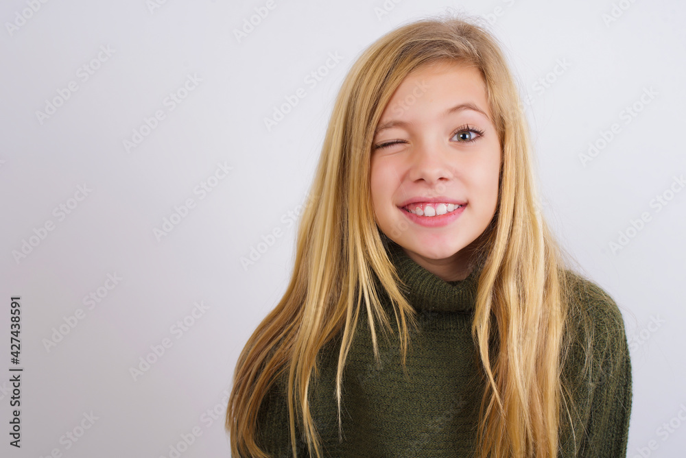 Coquettish Caucasian kid girl wearing green knitted sweater against white wall smiling happily, blinking at camera in a playful manner, flirting with you. Coquettish Caucasian kid girl wearing green knitted sweater against white wall smiling happily, blinking at camera in a playful manner, flirting with you.