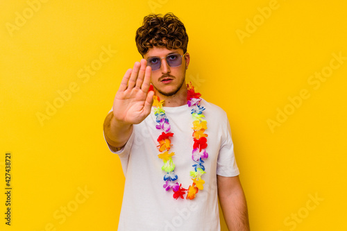 Young caucasian man wearing a Hawaiian party stuff isolated on yellow background standing with outstretched hand showing stop sign  preventing you.