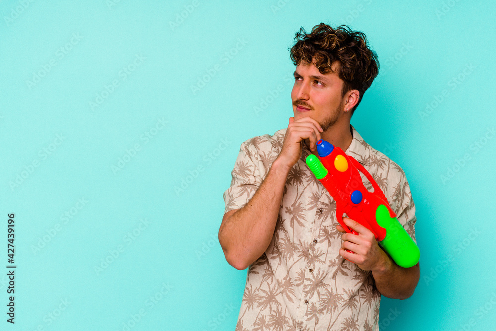 Young caucasian man holding a water gun isolated on blue background looking sideways with doubtful and skeptical expression. Young caucasian man holding a water gun isolated on blue background looking sideways with doubtful and skeptical expression.