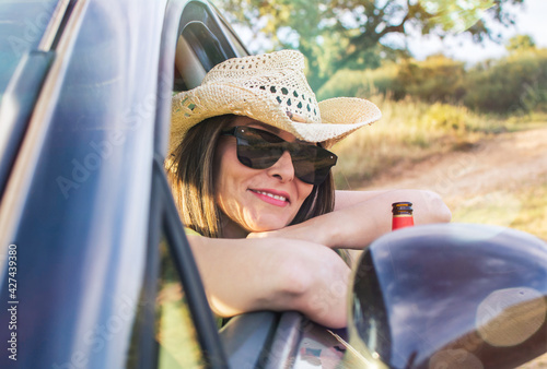 Young Woman Inside The Car Looking Out The Window With A Bottle Of Beer In Her Hand. She is wearing sunglasses and a hat and looking at the camera. Travel Concept