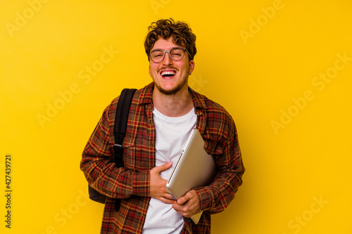 Young student caucasian man holding a laptop isolated on yellow background laughing and having fun.