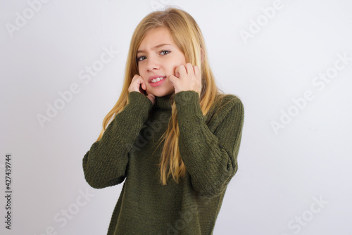 Caucasian kid girl wearing green knitted sweater against white wall covering ears with fingers with annoyed expression for the noise of loud music. Deaf concept.